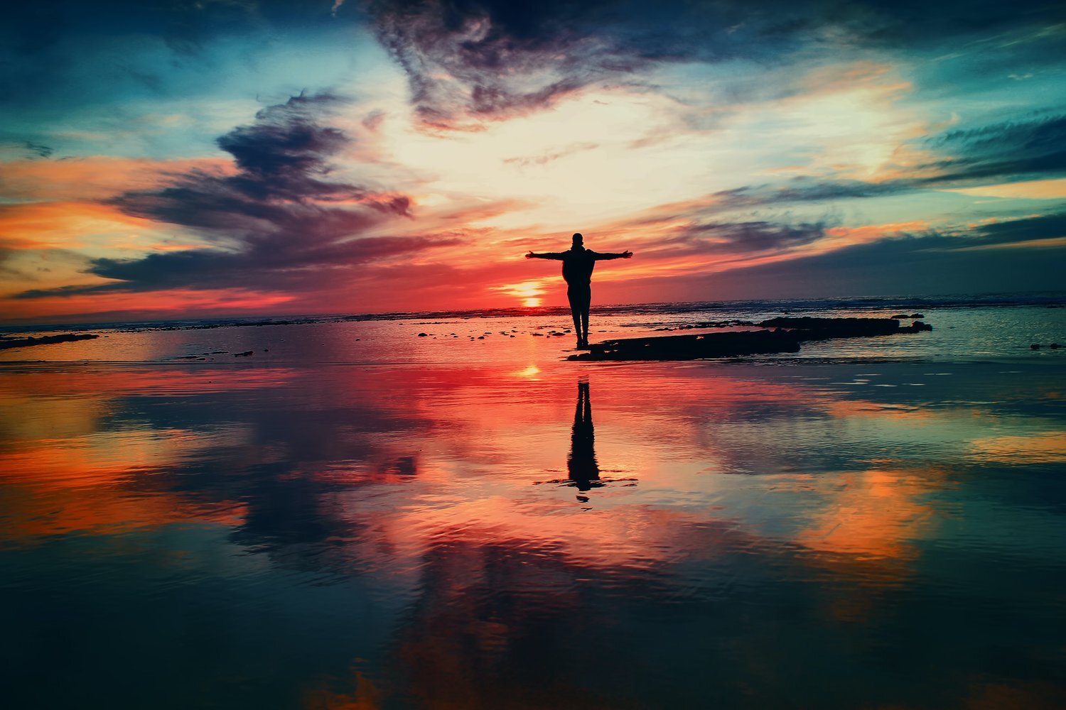 Silhouette of a person with outstretched arms standing by the ocean during a vibrant sunset, reflecting vivid colors on the water. The scene evokes freedom, peace, and connection with nature.